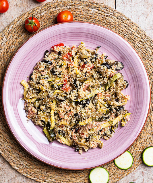 Cous cous con verdure grigliate e tonno servito su un piatto rosa in ceramica con quinoa e olive nere