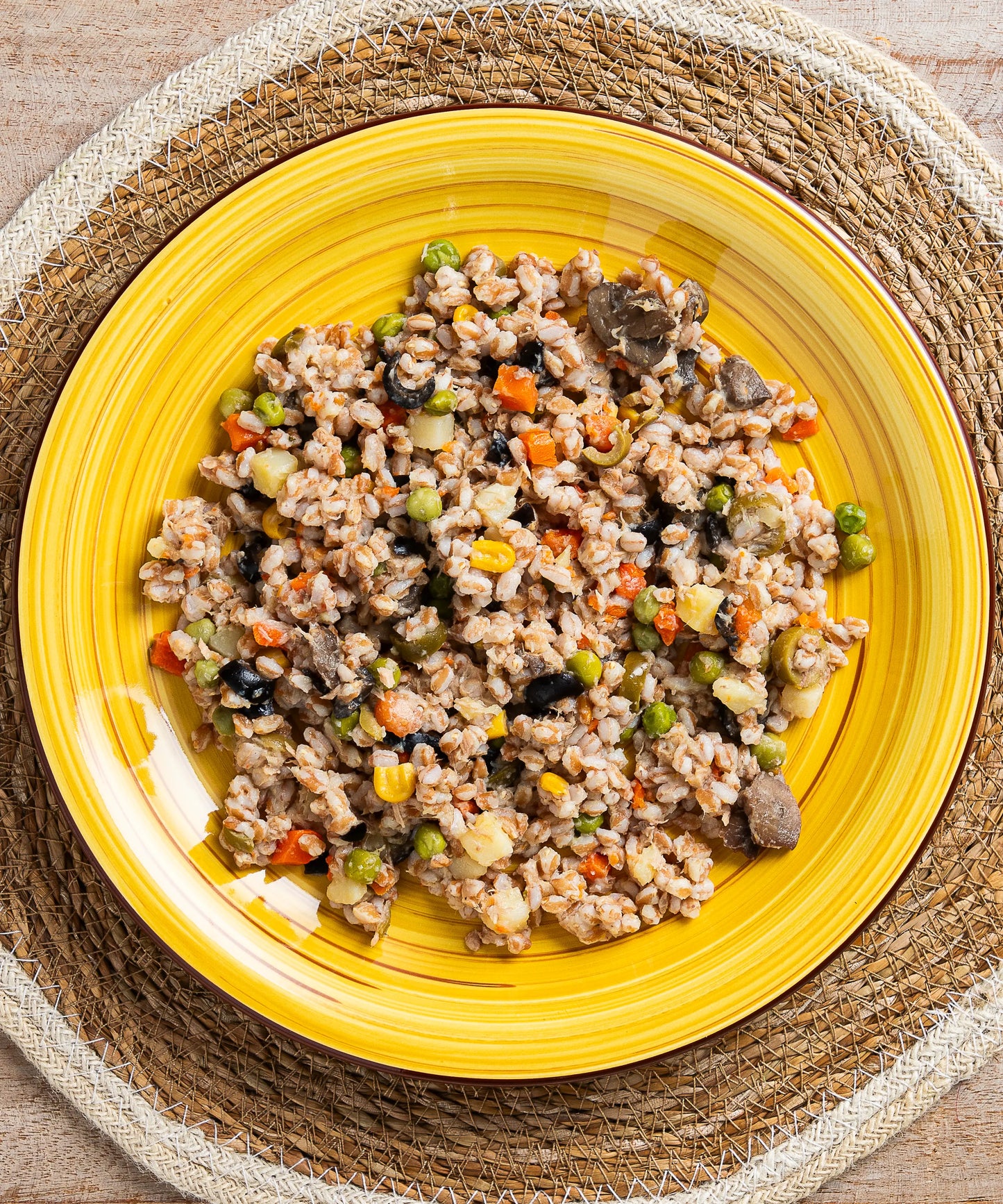 Insalata di farro con tonno funghi olive e mais servita su un piatto ceramico giallo vibrante con sottopiatto intrecciato
