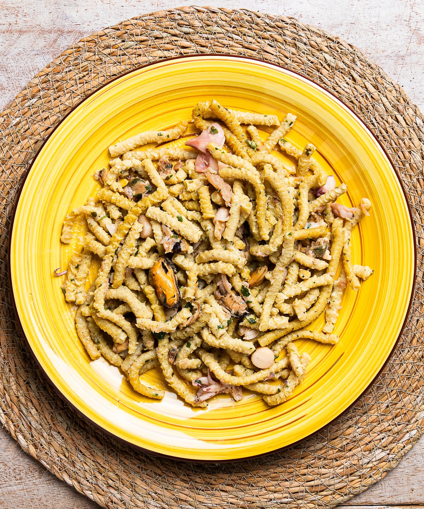 Un piatto giallo di ceramica contiene passatelli con frutti di mare, cozze, erbe e pezzi di prosciutto sale mare