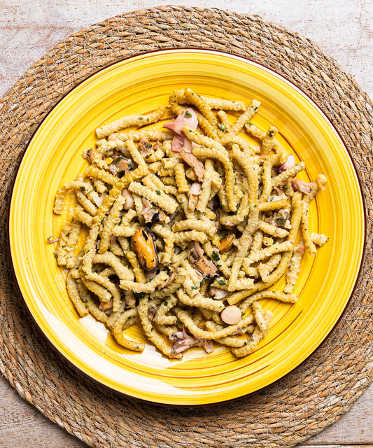 Un piatto giallo di ceramica contiene passatelli con frutti di mare, cozze, erbe e pezzi di prosciutto sale mare