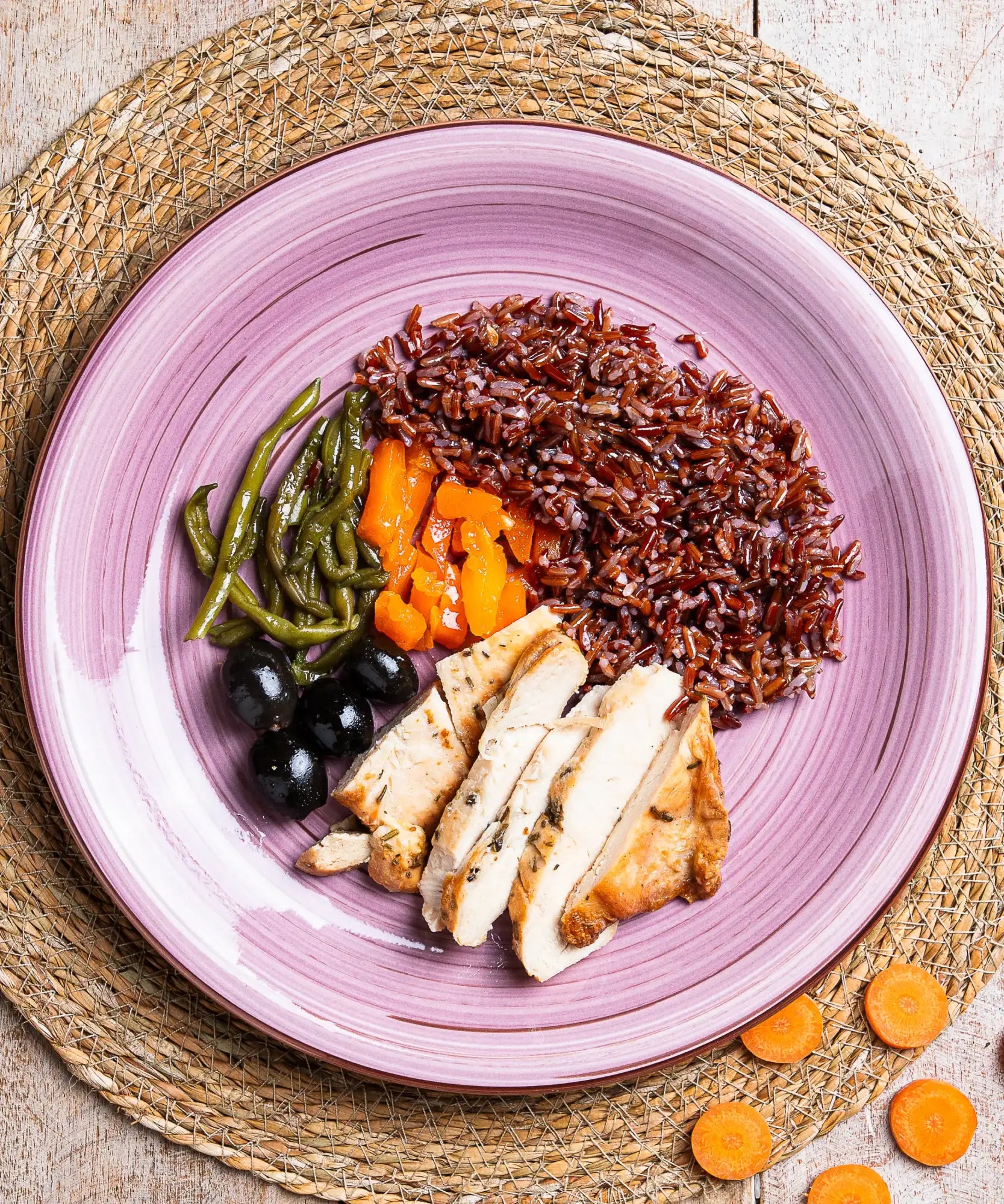 Un piatto di ceramica viola con motivi concentrici a rilievi contiene un servizio di riso integrale, fettine di petto di pollo, fagiolini verdi, carote arancioni e olive nere.