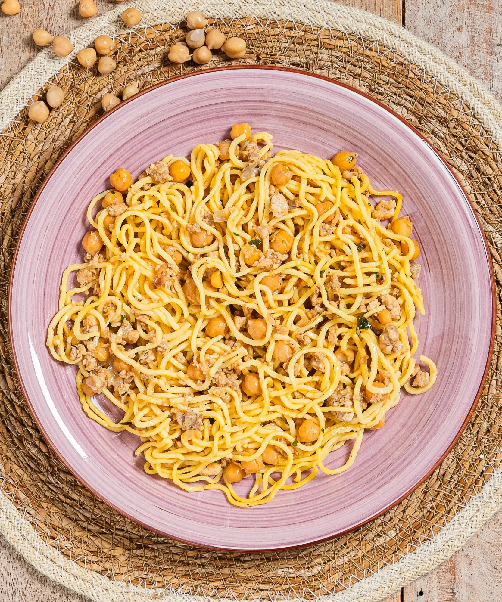 Un piatto di spaghetti gialli mescolati con ceci e carne macinata, serviti su un morbido piatto in ceramica rosa.