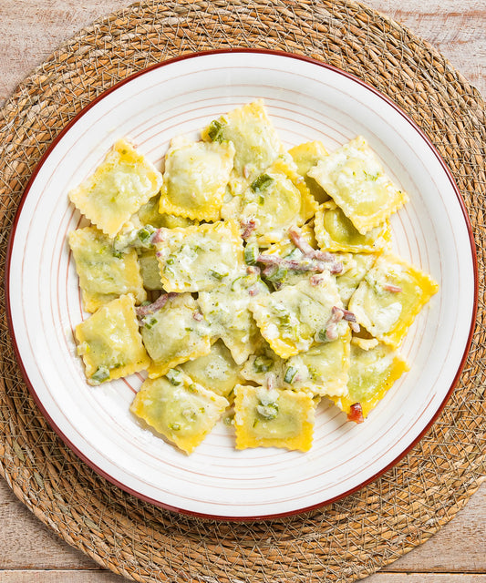 Una porzione di ravioli cremosi quadrati con speck alto adige igp e salsa verde con asparagi e pezzi di speck